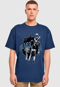 Marineblå bomulds T-shirt med grafisk tryk af en spiller og et ansigt, med fed skrift i blå og sort. Løs pasform, rund hals design.
