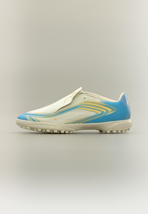 Zapatilla de fútbol baja en beige con patrones en remolino azul y amarillo y suela texturizada, mostrada en perfil lateral sobre fondo neutro.