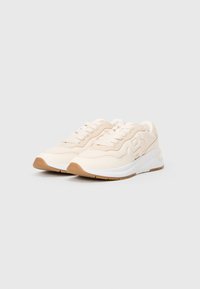 Beige és fehér sneakers pár, párnázott talppal és fűzőkkel, egymás mellett elhelyezve egy sima, világos háttér előtt.