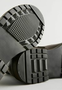 Las suelas de calzado negro presentan superficies texturizadas con patrones geométricos y sueltas anchas, diseñadas para ofrecer agarre y durabilidad.