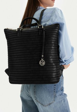 Mujer con camisa azul y vaqueros lleva una mochila grande negra con textura y un colgante circular plateado al hombro.