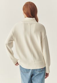 Crème-kleurige trui met een hoge kraag, geribbelde manchetten en zoom. Bevat een middenachter naad en is gecombineerd met lichtblauwe jeans.
