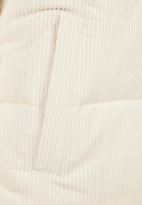 Gros plan d'un tissu côtelé de couleur crème présentant une couture horizontale et un patch rectangulaire en relief ou un détail en boucle.