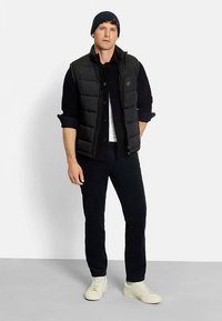 Schwarze Steppweste über einem dunkel gerippten Pullover, kombiniert mit marineblauen Hosen und weißen Sneakers. Eine marineblaue Mütze rundet das Outfit ab.