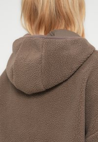 Άτομο με ξανθά μαλλιά που φοράει καφέ, υφασμάτινο φλις μπουφάν με κουκούλα, φαίνεται από πίσω σε απλό φόντο.