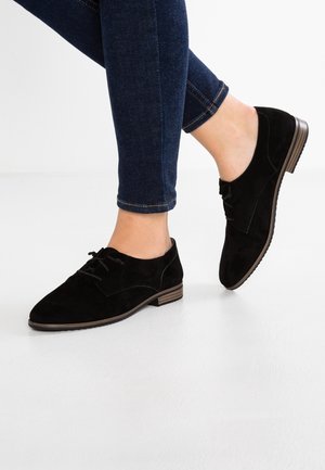 Person iført mørkeblå skinny jeans og sorte ruskind snørestøvler i oxford-stil, stående med den ene fod let løftet.