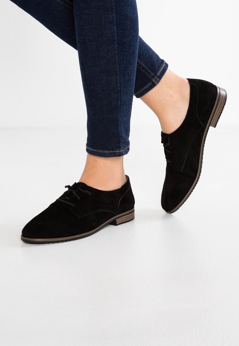Personne portant un jean slim bleu foncé et des chaussures Oxford en daim noir à lacets, debout avec un pied légèrement levé.