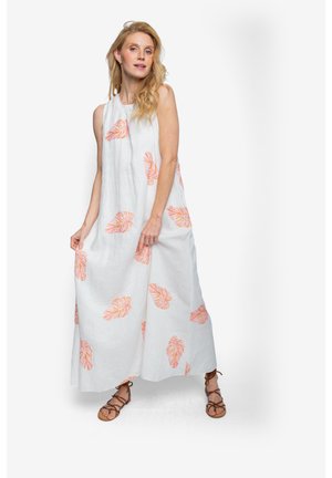 Kobieta z długimi blond włosami, ubrana w białą, bezrękawnikową sukienkę maxi z pomarańczowymi wzorami liści i brązowe sandały na sznurowadła.