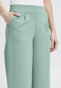 Lichtgroene wijde broek gemaakt van zachte stof met een hoge elastische tailleband en zijzakken; heeft subtiele plooien aan de voorkant.