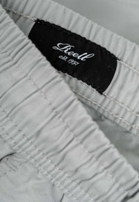 Pantalones cortos grises con cintura elástica y detalle de costura. Etiqueta negra con texto blanco que dice "Reell est. 1997." Textura de tela suave.