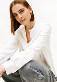 Blusa blanca de manga larga con cuello alto, con botones dorados y ligero fruncido en los hombros. Combinada con pantalones grises.