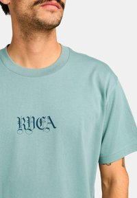 Light teal cotton t-shirt featuring a dark blue graphic logo "RVCA" with intricate lettering on the front, short sleeves, and a round neckline.