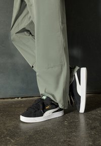 Czarne zamszowe sneakersy z białą gumową podeszwą, z czarnymi sznurowadłami, zieloną metką i białym paskiem po boku, zestawione z oliwkowo-zielonymi spodniami.