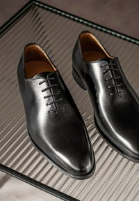 Zwarte leren dresschoenen met een spitse neus, voorzien van een gladde oppervlakte, vetersluiting en een minimalistische esthetiek op een gestreepte achtergrond.