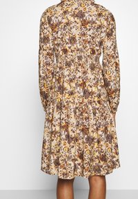 Pitkähihainen kukkakuosiset mekko, jossa on ruskeita, keltaisia ja kermanvärisiä sävyjä, vyötäröltä kiristettävä ja polviin ulottuva laskostettu hame.