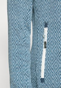 Blue patterned fleece jacket featuring a diamond design, textured fabric, and a white zippered pocket with a blue pull tab.