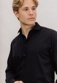 Svart langermet skjorte laget av glatt stoff, med spiss krage og syv knapper foran, med en smal passform.