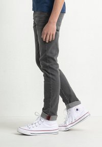 Jeans slim-fit grigi con tasche laterali, orli arrotolati e accenti rossi a contrasto, abbinati a sneakers alte bianche con dettagli blu.