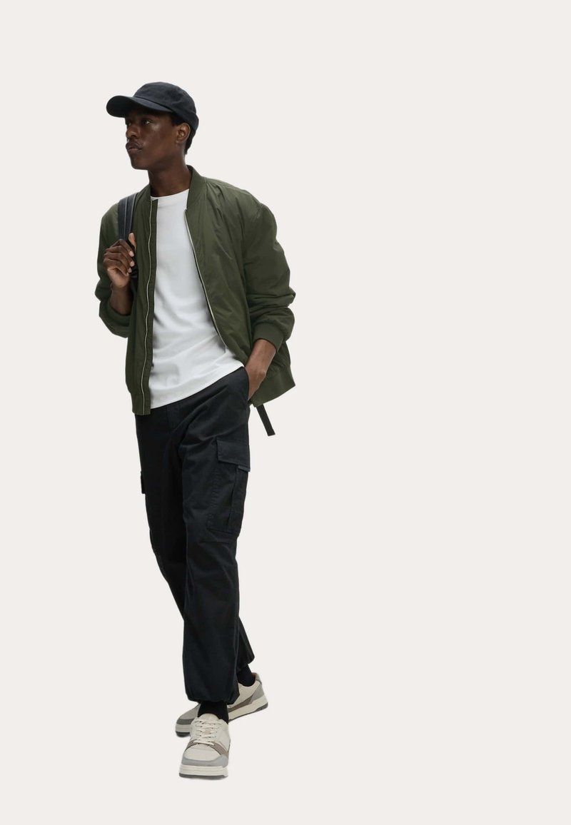 Giacca bomber verde oliva sopra una t-shirt bianca, pantaloni cargo neri e sneaker color crema. Cappellino nero, design slim-fit, texture del tessuto visibili.