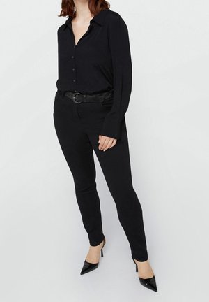 Vrouw die een zwarte blouse met knopen draagt, zwarte nauwsluitende broek met riem en zwarte spitse hoge hakken, staand tegen een effen achtergrond.