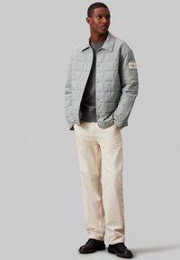 Hellgrüne gesteppte Jacke mit Druckknöpfen, getragen über einem dunkelgrauen Pullover, kombiniert mit cremefarbenen Hosen und schwarzen Schuhen.