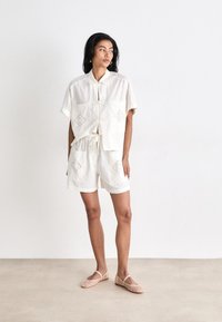 Completo in cotone bianco con apertura frontale, maniche corte e tasche ricamate. Abbinato a pantaloni corti bianchi e ballerine intrecciate rosa chiaro.