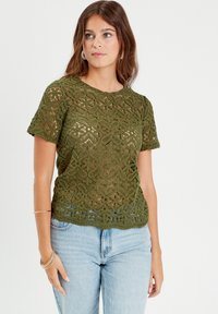 Haut en crochet vert olive avec un motif de dentelle florale, manches courtes et ourlet festonné, associé à un jean bleu clair.