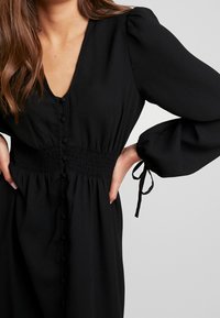 Schwarzes Kleid mit tiefem V-Ausschnitt, gerüschter Taille und Puffärmeln. Mit durchgehender Knopfleiste und Bindebändern an den Ärmeln. Glatte Stofftextur.