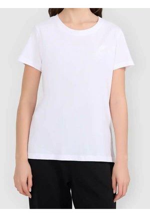 Kobieta ubrana w zwykły biały t-shirt z krótkim rękawem i okrągłym dekoltem oraz czarne spodnie, stoi na jasnym tle.