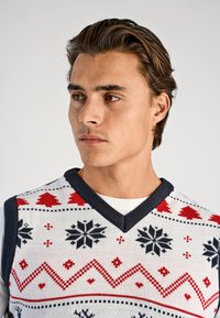 Gebreide sweater-vest met een witte basis en rode, zwarte en groene patronen, waaronder harten en sneeuwvlokken, met een marineblauwe V-halsafwerking.
