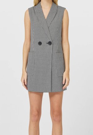 Vrouw draagt een mouwloze zwart-witte pied-de-poule blazerjurk met dubbele zwarte knopen, staand tegen een effen achtergrond.