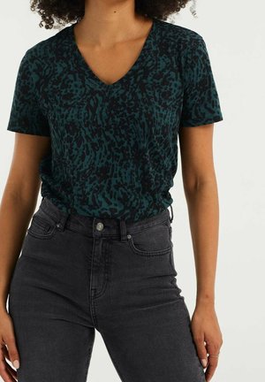 Kobieta ubrana w zieloną, ciemną, czarno-wzorzystą bluzkę z krótkim rękawem i dekoltem w serek, włożoną do wysokich czarnych jeansów.