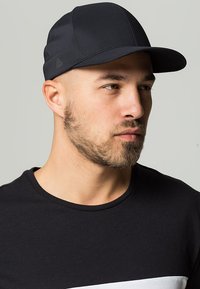 Schwarze Kappe mit gebogenem Schirm, aus einem glatten, atmungsaktiven Stoff. Verfügt über ein Logodetail an der Seite, kombiniert mit einem schwarzen Rundhals-T-Shirt.
