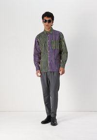 Camisa abotonada con rayas verticales en verde, púrpura y amarillo, con un bolsillo en el pecho. Combinada con pantalones grises y zapatos negros.