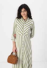 Rochie rayée verde și alb cu mâneci lungi și închidere cu butoane, asociată cu o geantă mică maro cu textură matlasată.