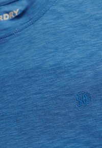 Zbliżenie na niebieską tkaninę o fakturze, z subtelnym wyszywanym logo i częściowym białym nadrukiem tekstu w lewym górnym rogu.