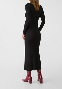 Robe noire côtelée à manches longues avec une coupe ajustée et une fente latérale, associée à des bottines bordeaux brillantes à talon carré.