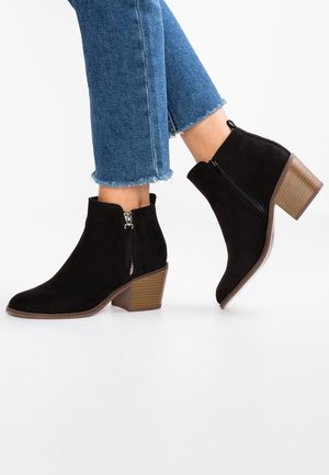 Sorte ankelstøvler i ruskind med sidelommer og blokhæle, båret med korte blå jeans på en hvid baggrund.