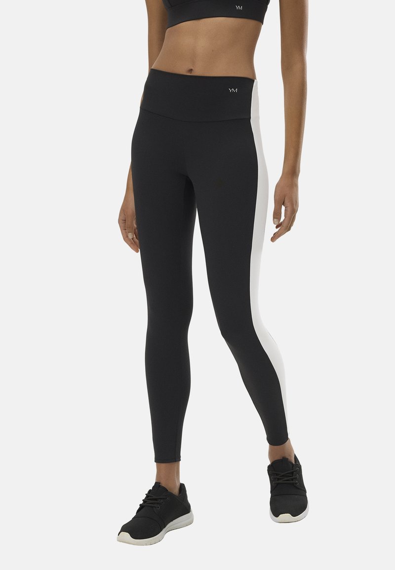 Mujer que lleva leggings de cintura alta en blanco y negro, combinados con un sujetador deportivo negro y zapatillas deportivas negras con suelas blancas.