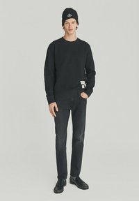 Felpa nera con collo a giro e una piccola etichetta sull'orlo, abbinata a jeans neri slim-fit e scarpe nere, completata da un berretto nero.