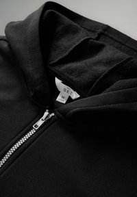 Schwarze Zip-Hoodie mit weicher Textur, hohem Kragen und sichtbarem silbernen Reißverschluss. Das Etikett lautet "THE SET" und die Größe ist "M."