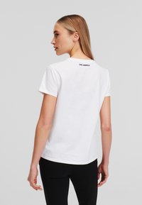 Hvit bomullst-skjorte med rund hals og korte ermer, med en svart "KARL LAGERFELD" logo bak på nakken. Enkelt design.