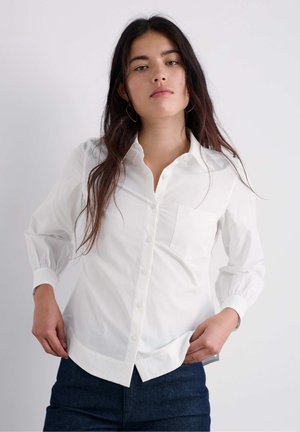 Ženska z dolgimi temnimi lasmi, ki nosi zlate obročaste uhane, belo srajco z gumbi in temno modre kavbojke, stoji ob svetli enobarvni podlagi.