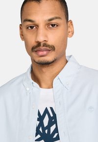Uomo con capelli corti e barba curata indossa una camicia azzurro chiaro sopra una maglietta bianca con grafica e design geometrico blu navy.
