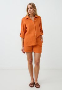 Oranje, gestructureerd shirt met een knoopsluiting aan de voorkant en opgerolde mouwen, gecombineerd met bijpassende shorts. Beide stukken hebben een relaxte pasvorm.
