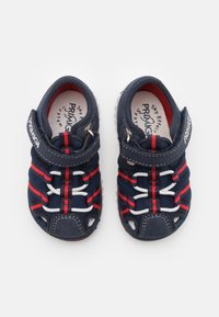 Marinblå barnskor med röda och vita elastiska snören, öppen tå och VELCRO-rem. Slät ovandel i material med gummisula.