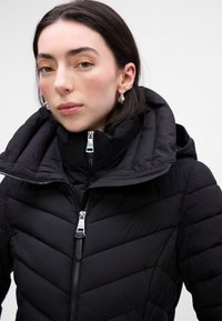 Giacca a vento nera con colletto alto, struttura trapuntata e dettagli in zip argentati. Presenta un cappuccio staccabile e un taglio moderno.