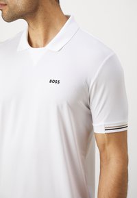 Λευκό πόλο μπλουζάκι από υφασμάτινη υφή, με αιχμηρό γιακά, μικρό μαύρο λογότυπο "BOSS" και ριγέ λεπτομέρειες στα μανίκια.