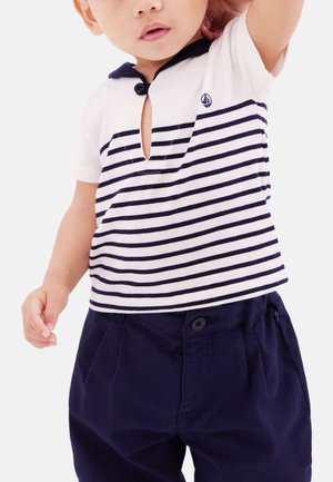 Maluch ubrany w biało-granatową koszulkę w paski z małym logo żaglówki i ciemnogranatowe spodnie, unoszący jedną rękę.