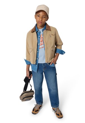 Beige jas met een bruine kraag, blauw denim overhemd, grafisch T-shirt, blauwe jeans, sportieve schoenen met beige accenten en een beige pet. Houdt een kleine tas vast.